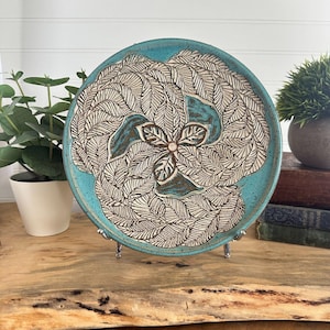 May include: A round ceramic plate featuring a detailed, carved leaf pattern arranged in a circular, swirling design. The surface displays a vibrant turquoise glaze with dark, recessed lines highlighting the intricate texture of the foliage.
