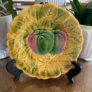 May include: A decorative ceramic plate with a leaf design in yellow, green, and pink. The plate features two stylized fruits in the center and is displayed on a black stand. The plate has a scalloped edge.