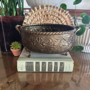 May include: An antique bronze-colored metal planter with ornate floral detailing and small feet. The planter has two handles and sits atop a vintage book. A woven fan and potted plants are in the background.