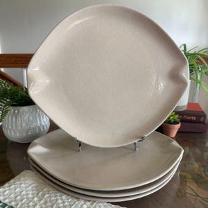 May include: A set of three stacked ceramic plates in a light beige color. The plates feature a unique square shape with rounded, slightly indented sides and a smooth, glossy finish. The top plate is displayed upright on a small metal stand.