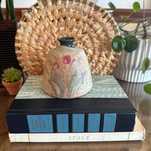 May include: A small, bell-shaped ceramic vase with a speckled beige finish, featuring hand-painted pink floral motifs and a dark blue rim. The vase sits atop a stack of two books, with the bottom book displaying the word ITALY on its spine.