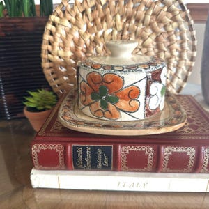 May include: A ceramic butter dish with a lid featuring a hand-painted floral pattern in shades of orange, green, and brown on a cream background. The dish has a square base with rounded corners and a matching lid with a small knob handle on top.