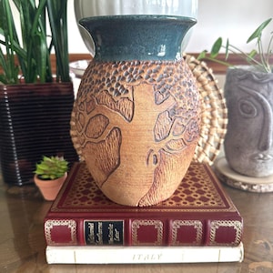 May include: A handcrafted ceramic vase with a textured, tree-like design in shades of brown and teal. The vase is displayed on a stack of books, including one with the title "The Scarlet Letter". The vase is a decorative item.