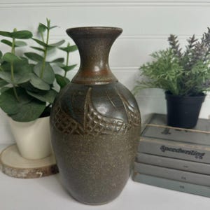 May include: A dark brown ceramic vase with a flared rim and a textured, carved grid pattern around the upper body. The surface has a mottled, earthy finish with subtle blue and copper-toned highlights throughout the glaze.