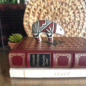 May include: A ceramic bear figurine featuring a white base with geometric patterns in black and terracotta red. The bear is positioned on top of a red book titled The Scarlet Letter by Nathaniel Hawthorne, which rests on a white book labeled ITALY.