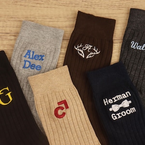 Puede incluir: Calcetines acanalados surtidos en negro, gris, marrón y beige, cada uno bordado con texto o diseños personalizados en varios colores. Los calcetines presentan nombres, iniciales y un diseño de pajarita, ideales para regalos personalizados.