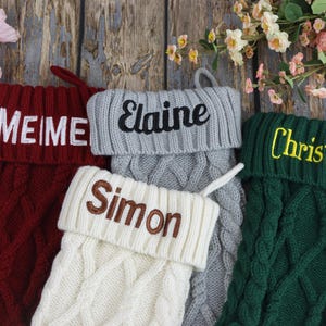 May include: Four cable-knit Christmas stockings in red, grey, white, and green, each with a different name embroidered in a contrasting colour: "MEIME", "Elaine", "Simon", and "Christina". The stockings are displayed on a wooden surface with floral accents.