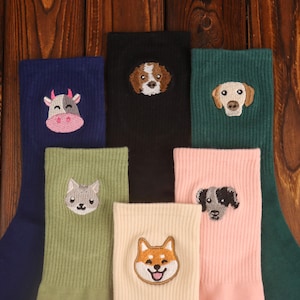 Könnte beinhalten: Eine Sammlung bunter Socken, jede mit einem gestickten Tiergesicht. Die Socken sind in verschiedenen Farben erhältlich, darunter Marineblau, Schwarz, Grün und Rosa. Zu den gestickten Tieren gehören eine Kuh, ein Hund, eine Katze und ein Shiba Inu.