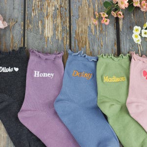 Può includere: Cinque paia di calzini a coste in vari colori: antracite, lavanda, blu, verde e rosa. Ogni calzino ha un bordo superiore arricciato e testo ricamato: "Olivia", "Honey", "Daisy", "Madison" e un fiocco rosa.