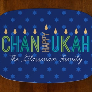 Peut inclure: Un plat de service bleu ovale avec des étoiles bleues. Les mots "Happy Chanukah" sont écrits en vert, bleu et or, avec "The Glassman Family" en dessous. Le plat est sur une surface en bois.