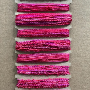 May include: A collection of vibrant pink yarn samples, each wound on a cardboard holder. The yarns vary in texture, from smooth to heavily textured, and display a range of pink hues, including fuchsia and magenta. Ideal for crafting and textile projects.