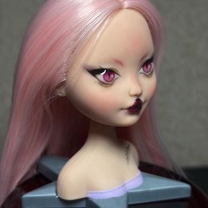 May include: A doll head with long, light pink hair and detailed facial features. The doll has pink eyes, dark lipstick, and is mounted on a gray star-shaped base. The doll's skin is pale with blush.
