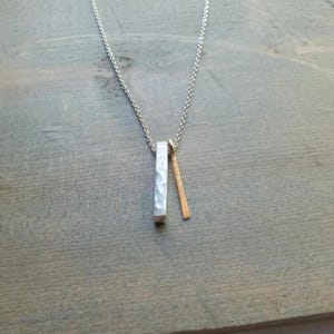 May include: A silver necklace with a hammered rectangular pendant and a smaller gold bar pendant. The pendants hang from a delicate silver chain. The necklace is displayed on a wooden surface.
