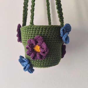 May include: A green crocheted hanging planter with purple and blue flower accents.
