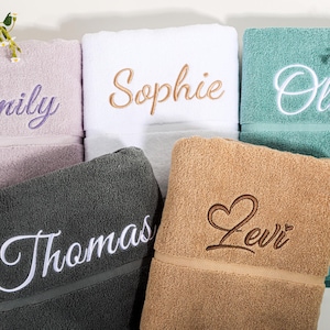 May include: A selection of embroidered bath towels in shades of lavender, white, teal, charcoal, and tan. Each towel features a name embroidered in a coordinating colour, including "Emily," "Sophie," "Olivia," "Thomas," and "Levi."