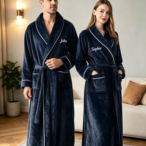 以下が含まれることがあります： ショールカラー、袖口、ポケットに白いパイピングが施されたネイビーブルーのプラッシュバスローブ2着。一方の左胸には「John」、もう一方には「Sophie」という名前が白い筆記体で刺繍されています。