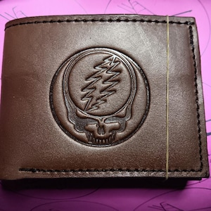 May include: Brown leather wallet with a debossed Steal Your Face logo. The wallet is rectangular and has a stitched border. A thin rubber band is wrapped around the wallet.