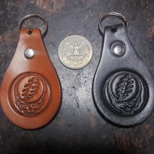 May include: Two leather keychains, one brown and one black, each featuring a Steal Your Face design. The keychains have a silver metal ring and a silver rivet. A quarter is placed between the keychains.