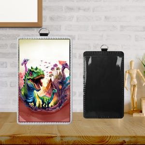 Può includere: Una stampa artistica incorniciata con una colorata scena di dinosauri con un T-Rex, montagne e altri dinosauri. La cornice è bianca con un anello argentato in alto. Accanto, un porta badge nero in similpelle.