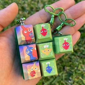 Puede incluir: Varios llaveros cuadrados con personajes Pikmin coloridos. Cada llavero tiene un borde verde y un cierre de metal. Los Pikmin se representan en varios colores, sobre un fondo de paisaje.