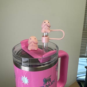 May include: Pink stainless steel travel mug with a handle and a lid. The mug has a white star pattern and the word "STANLEY" printed on the side. The lid has a straw holder and two pink silicone figures on top.