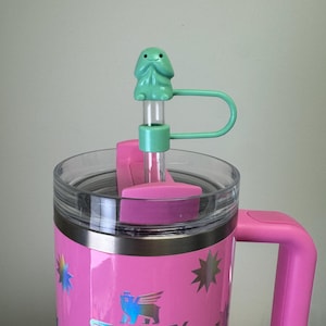 May include: A pink stainless steel travel mug with a black handle and a lid. The mug has a white logo that says "STANLEY" and a holographic star design. The lid has a clear plastic top and a green plastic straw with a green frog-shaped topper.