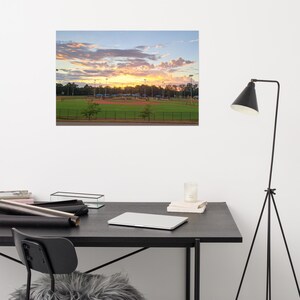 Könnte beinhalten: Ein Druck eines Baseballfeldes bei Sonnenuntergang. Der Himmel ist mit leuchtenden Orange-, Gelb- und Rosatönen und flauschigen Wolken gefüllt. Das Feld hat ein rotes Lehm-Infield, grünes Gras und einen schwarzen Zaun. Bäume und Lichter sind sichtbar.