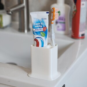 May include: A white toothbrush holder containing a tube of Halson "fresh & minty" toothpaste and an orange and white toothbrush. The toothpaste tube features blue and white swirls and a green mint leaf graphic. The holder is on a white sink.