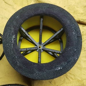 May include: A close-up of a black foam tire with a yellow interior and a black, star-shaped spoke design. The tire is likely for a small vehicle or model, with a textured surface.