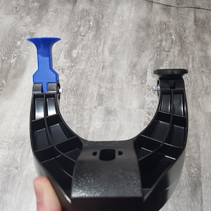 Può includere: Staffa a forma di U in plastica nera con un pezzo di plastica blu attaccato nella parte superiore. La staffa ha supporti interni e una base piatta. Una mano tiene la parte inferiore della staffa.