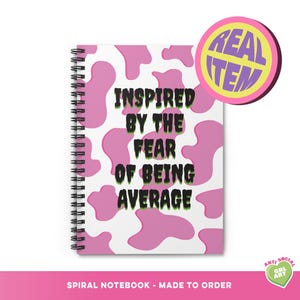Peut inclure: Cahier à spirales avec un motif de vache rose et blanc. La couverture porte l'inscription "INSPIRED BY THE FEAR OF BEING AVERAGE" en noir et vert. Un autocollant rond avec les mots "REAL ITEM" est dans le coin supérieur droit.