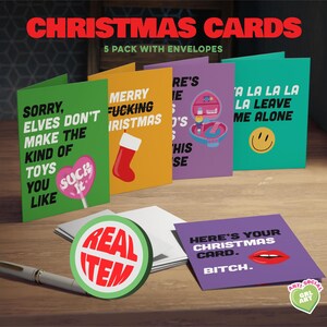 May include: A set of five Christmas cards with envelopes, each featuring bold text and graphics. The cards have various colors, including green, yellow, purple, and teal. One card has a smiley face, and another has a heart-shaped lollipop.