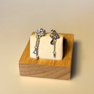 Puede incluir: Un par de pendientes plateados con forma de nube con cadenas colgantes y gemas azules y moradas en forma de lágrima. Los pendientes se exhiben sobre un pequeño bloque blanco cuadrado sobre un soporte de madera.