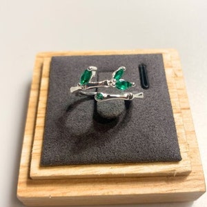 May include: A silver ring featuring a delicate branch design with emerald green leaves. The ring is presented in a wooden ring box with a dark grey velvet lining.