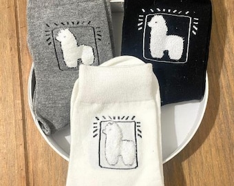 Puede incluir: Tres pares de calcetines con un diseño de alpaca blanco en un marco negro. Un par es gris, uno es negro y uno es blanco.