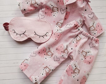 Puede incluir: Un conjunto de pijama rosa con un estampado de búhos y corazones, que incluye una camisa con cuello, pantalones y una máscara para dormir a juego. La máscara para dormir tiene pestañas bordadas. El conjunto está diseñado para una muñeca o un niño pequeño.