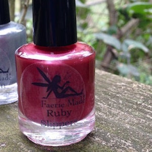 May include: A bottle of Faerie Made Ruby Slippers nail polish. The nail polish is a deep red color with a black cap. The label features a silhouette of a fairy. The bottle is on a wooden surface.