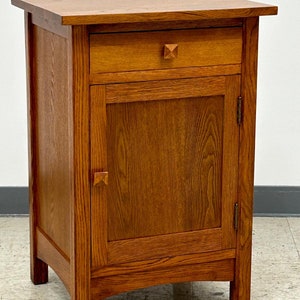 May include: A wooden nightstand with a single drawer and a cabinet door. The nightstand is made of oak and has a warm, reddish-brown finish. The drawer and door feature geometric wooden knobs. The nightstand has a rectangular top and sits on four sturdy legs.