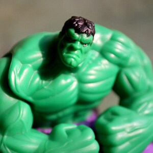 May include: A green plastic action figure of the Hulk, a superhero with green skin and a muscular physique. The figure is posed with its fists clenched and its arms crossed. The figure is wearing purple pants.