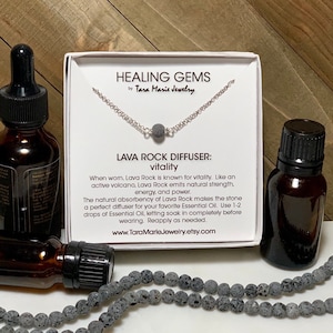 May include: A silver chain necklace with a lava rock diffuser pendant. The necklace comes in a small white box with the text "HEALING GEMS by Tara Marie Jewelry" and "LAVA ROCK DIFFUSER: vitality". The text explains that lava rock is known for vitality and can be used as a diffuser for essential oils. The box is next to two small bottles of essential oil.