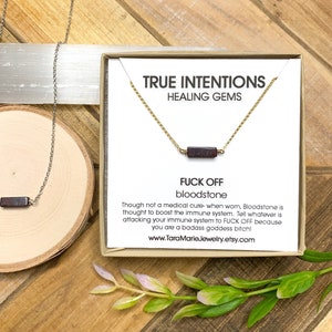 May include: A gold chain necklace with a dark red bloodstone pendant. The necklace comes in a small box with the text "TRUE INTENTIONS HEALING GEMS" and "FUCK OFF bloodstone" with a description about the healing properties of the stone.