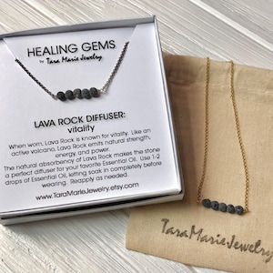May include: A gold chain necklace with a small row of black lava rock beads. The necklace comes in a white box with the text "HEALING GEMS by Tara Marie Jewelry" and a description of the lava rock diffuser. The necklace is also shown in a small brown pouch with the text "Tara Marie Jewelry".