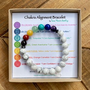 May include: A chakra alignment bracelet with white howlite beads and colorful gemstone beads representing the seven chakras. The bracelet is in a small box with a card that lists the chakras and their corresponding affirmations. The card reads "Chakra Alignment Bracelet by Tara Marie Jewelry".