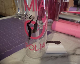 Può includere: Una bottiglia d'acqua in plastica trasparente con un tappo argentato e un design rosa. Il design include il nome "MIA" e una silhouette di una ginnasta con il testo "POL14".