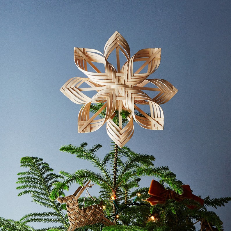 May include: A woven straw star ornament sits atop a green Christmas tree. The star is a traditional Scandinavian design, with a light brown colour and a delicate, intricate pattern.