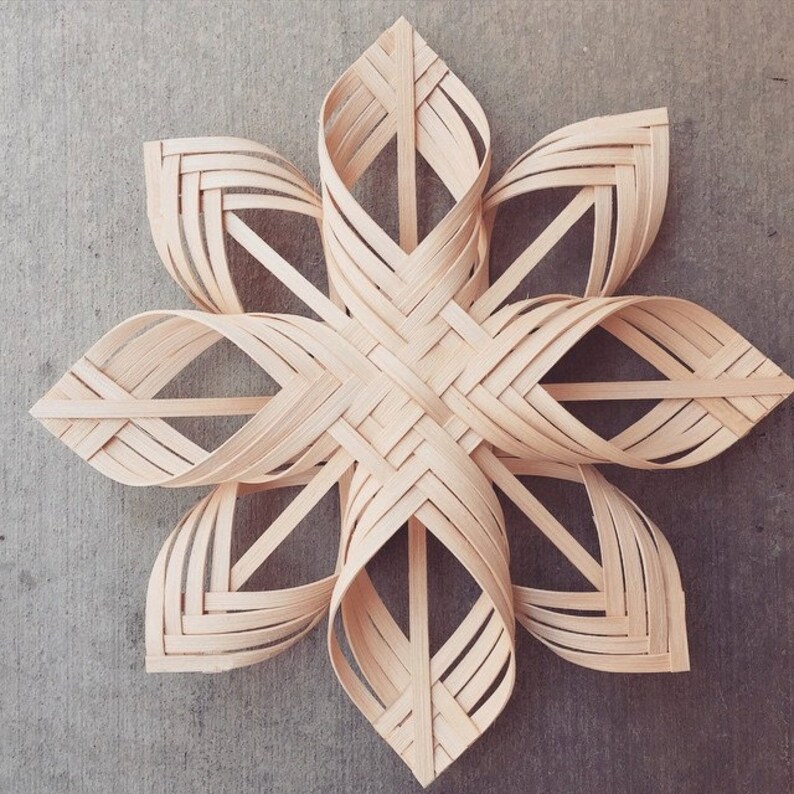 May include: A woven wooden star ornament in a light brown colour. The star is made of thin strips of wood that are woven together to create an intricate pattern.
