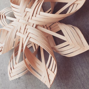 May include: A woven star-shaped ornament made of light brown wood. The star is made of thin strips of wood that are woven together to create a three-dimensional shape.