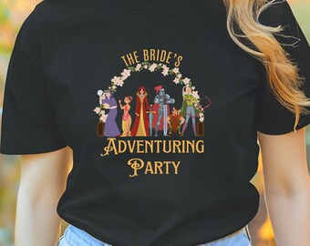 May include: Black t-shirt with the text "THE BRIDE'S ADVENTURING PARTY" in a floral wreath design. The shirt features a group of cartoon characters. The model is wearing blue jeans and glasses, standing in a field of flowers.