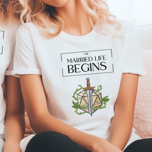 May include: Two people wearing white t-shirts with a graphic of a sword and a dice with the text "The Married Life Begins".