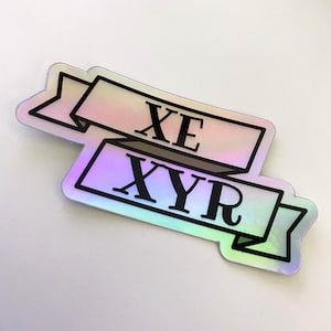 May include: Holographic sticker featuring two overlapping banners with the text "XE" and "XYR" in black. The banners have a pearlescent, iridescent finish with a black border. The sticker is on a white background.